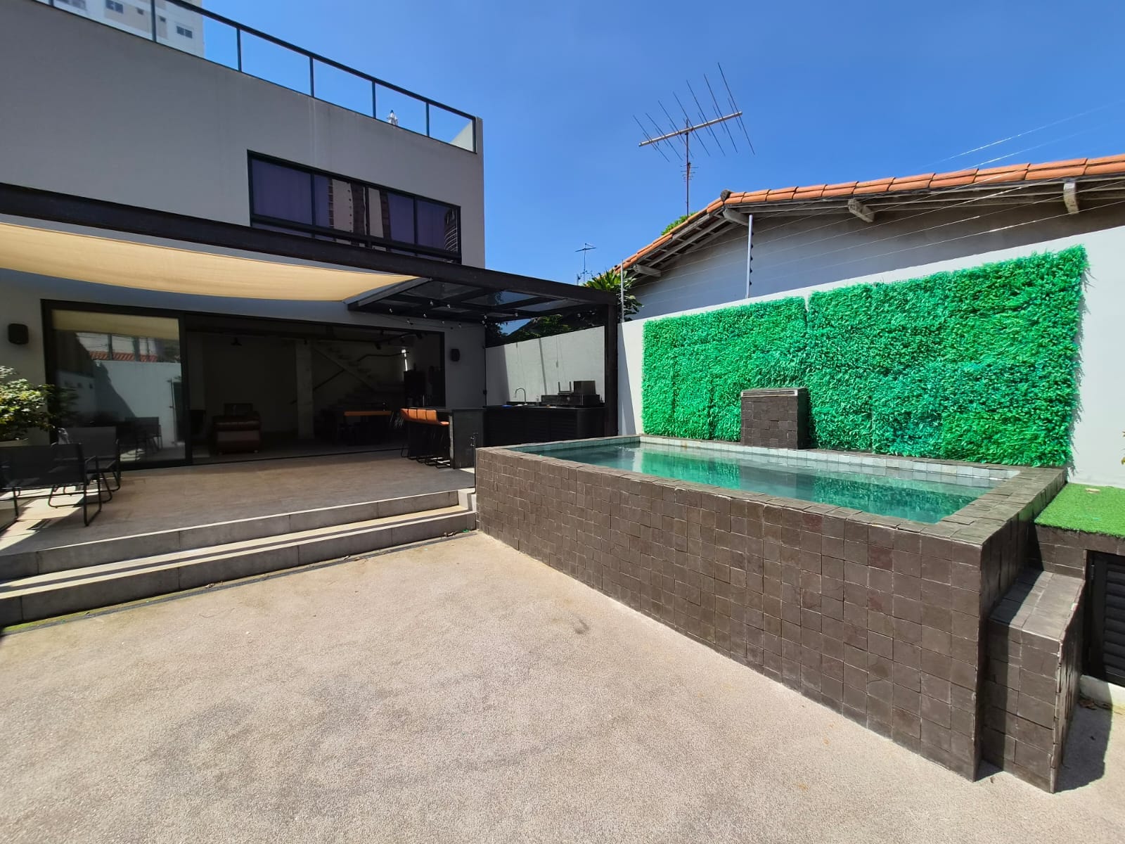 Área externa com piscina elevada, parede verde, cobertura de vidro e vista para a sala