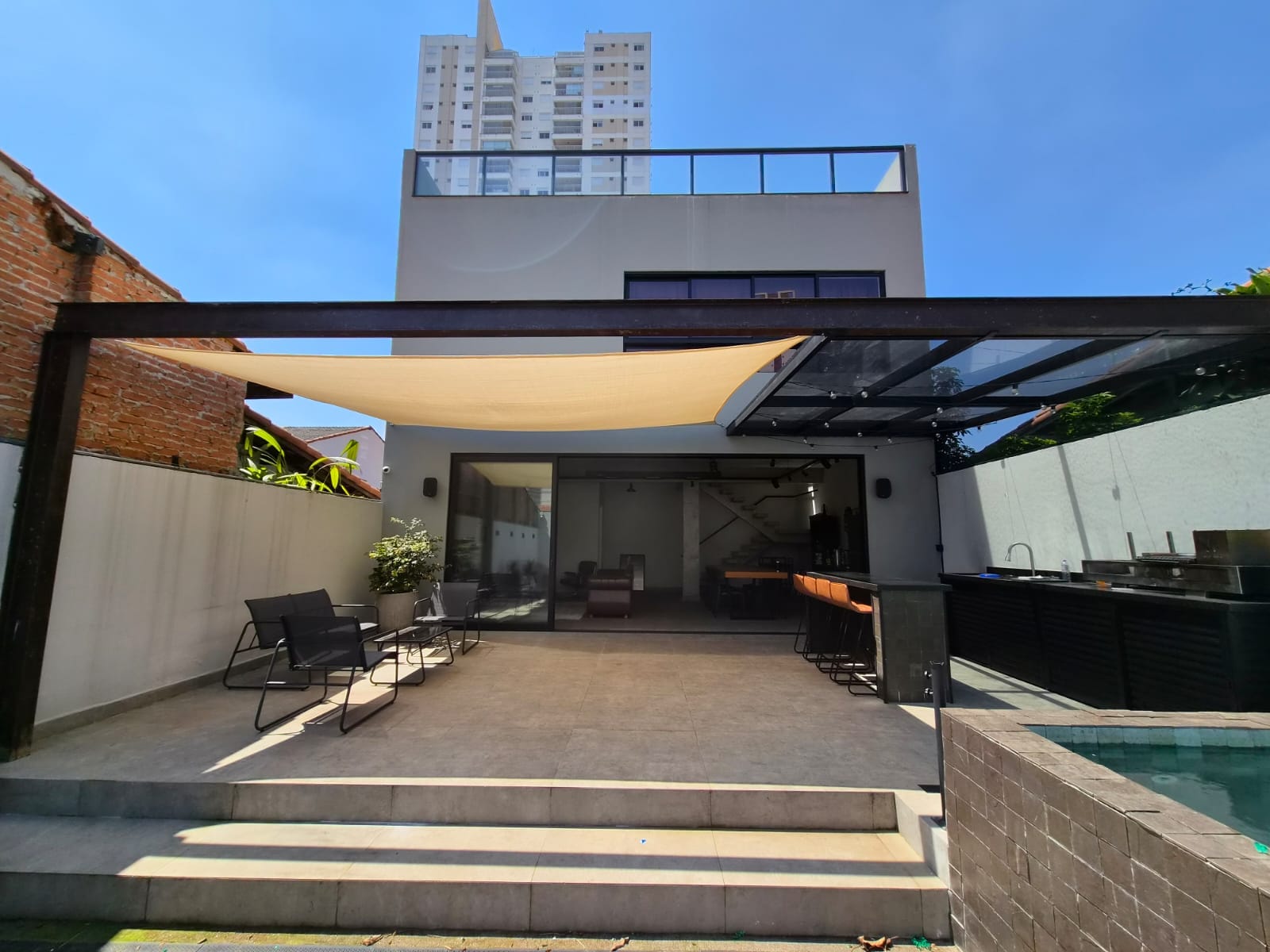 Casa vista do pátio: pergolado metálico, vela de sombreamento, portas de vidro abertas e área gourmet ao lado da piscina