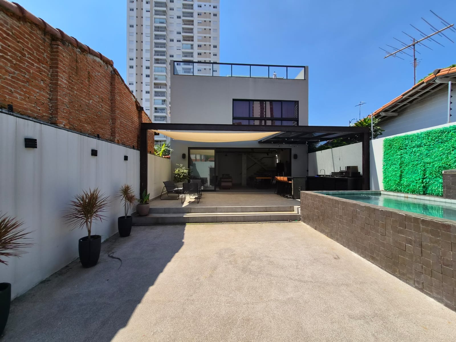 Vista geral do Brooklin 46: pátio de concreto, pergolado metálico com vela de sombreamento, muro de tijolos aparentes e piscina