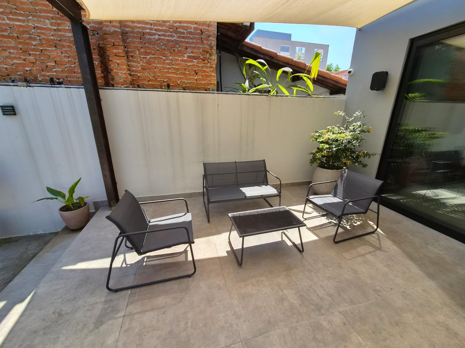 Estar externo sob a vela de sombreamento, com poltronas metálicas, vasos de plantas e muro de tijolos aparentes