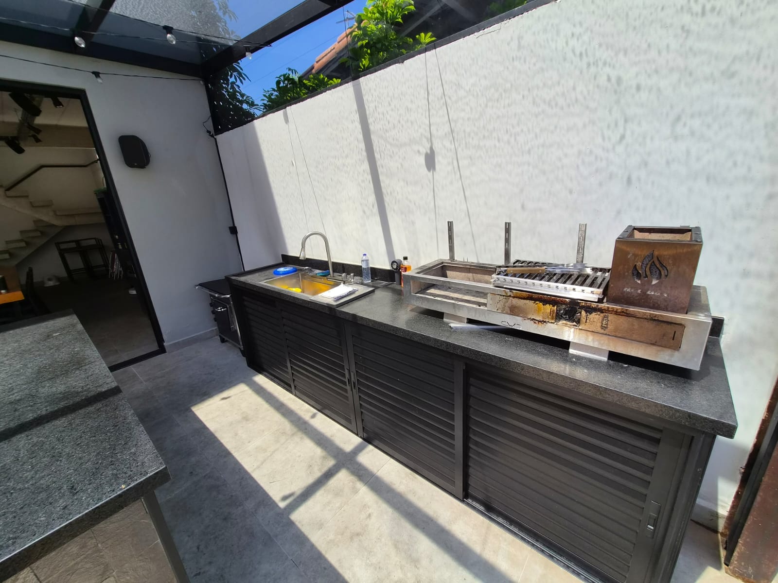 Bancada da área gourmet com pia, churrasqueira e armários pretos, sob cobertura de vidro
