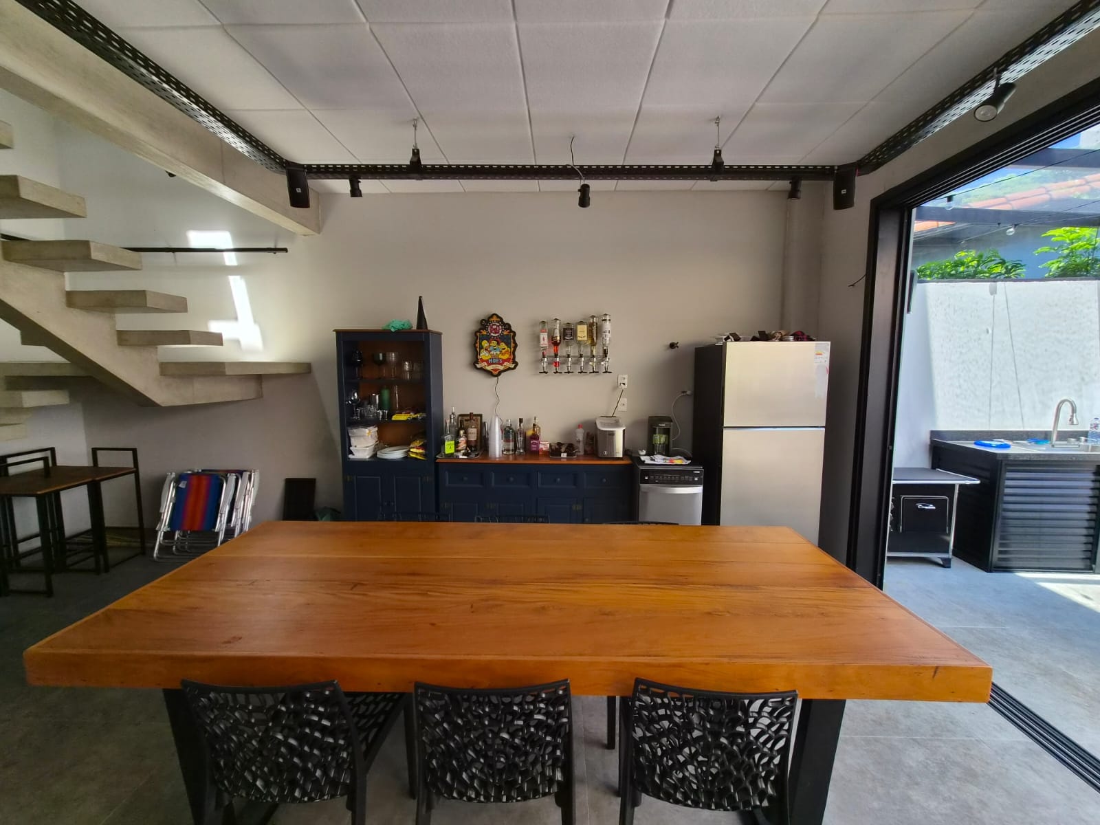 Mesa de jantar de madeira maciça com cadeiras pretas, escada de concreto e cristaleira azul ao fundo