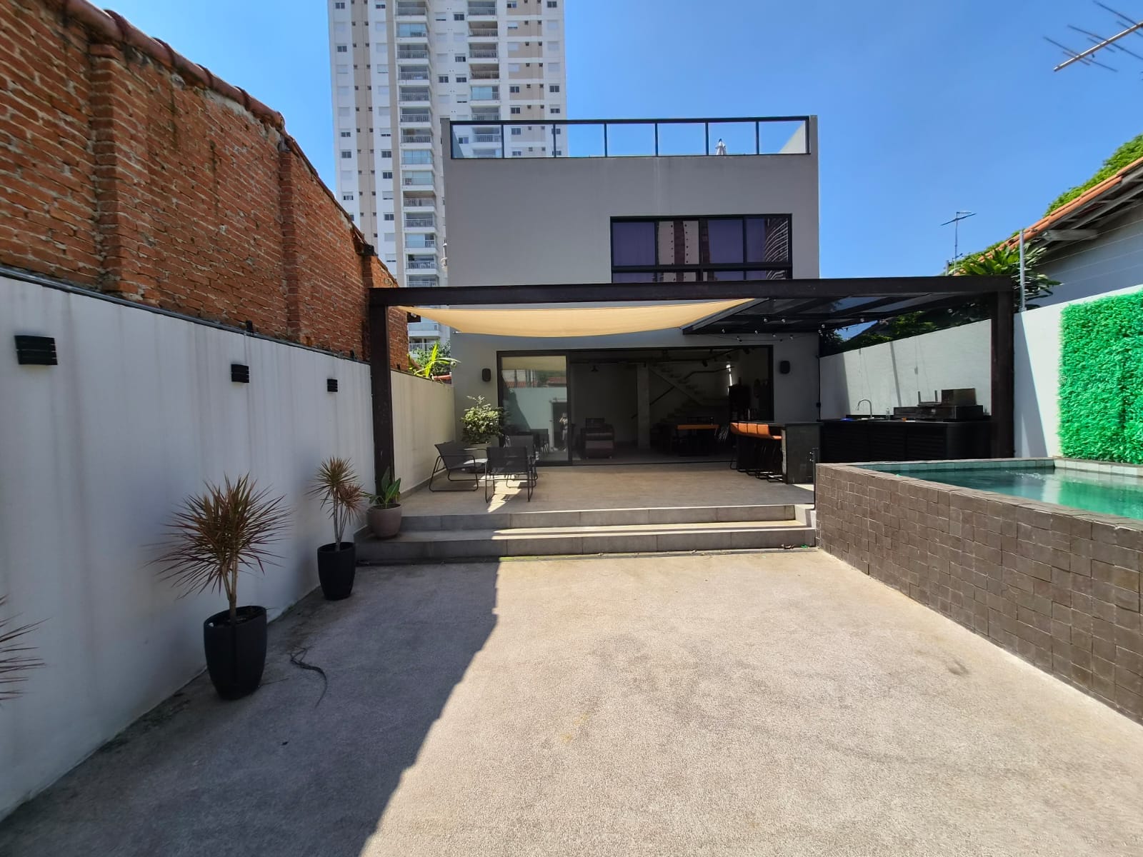 Pátio com muro de tijolos aparentes, vasos de plantas, pergolado e piscina