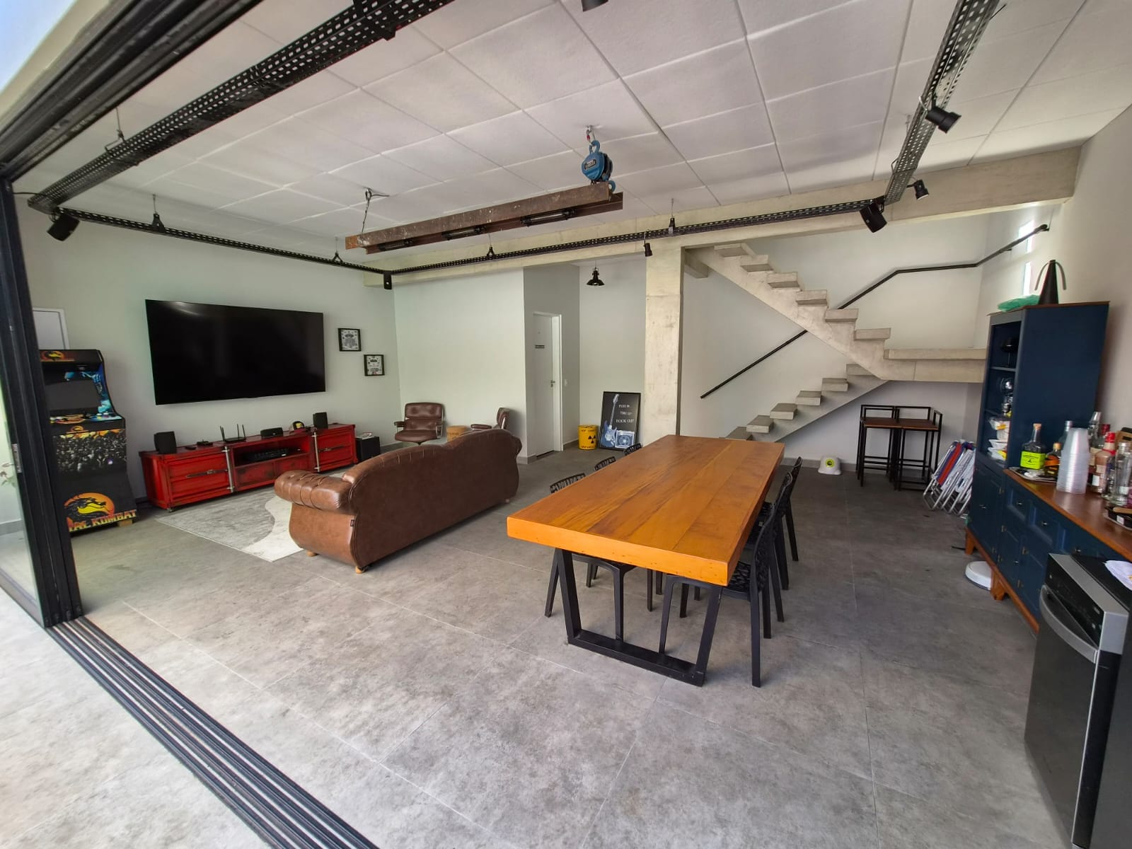 Sala ampla com piso de concreto, escada de concreto, trilhos de iluminação metálicos, mesa grande de madeira e sofá de couro