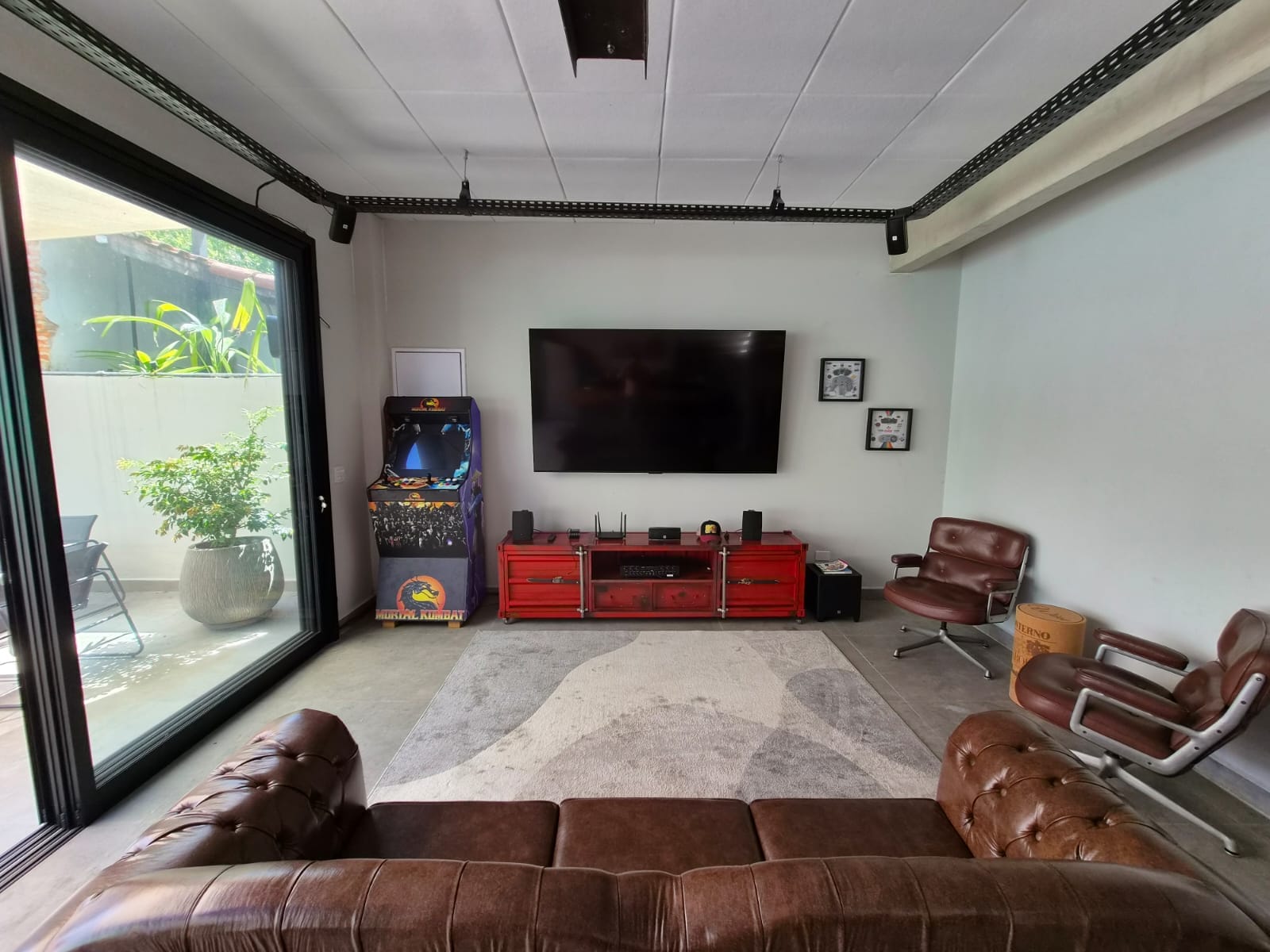Sala de estar com sofá de couro, poltronas, TV grande, rack vermelho estilo contêiner e fliperama