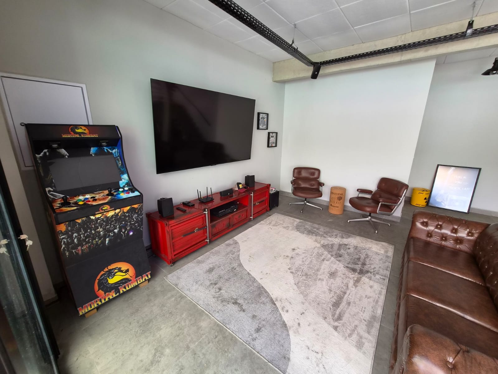 Sala de estar com poltronas de couro, TV, fliperama e tapete sobre piso de concreto