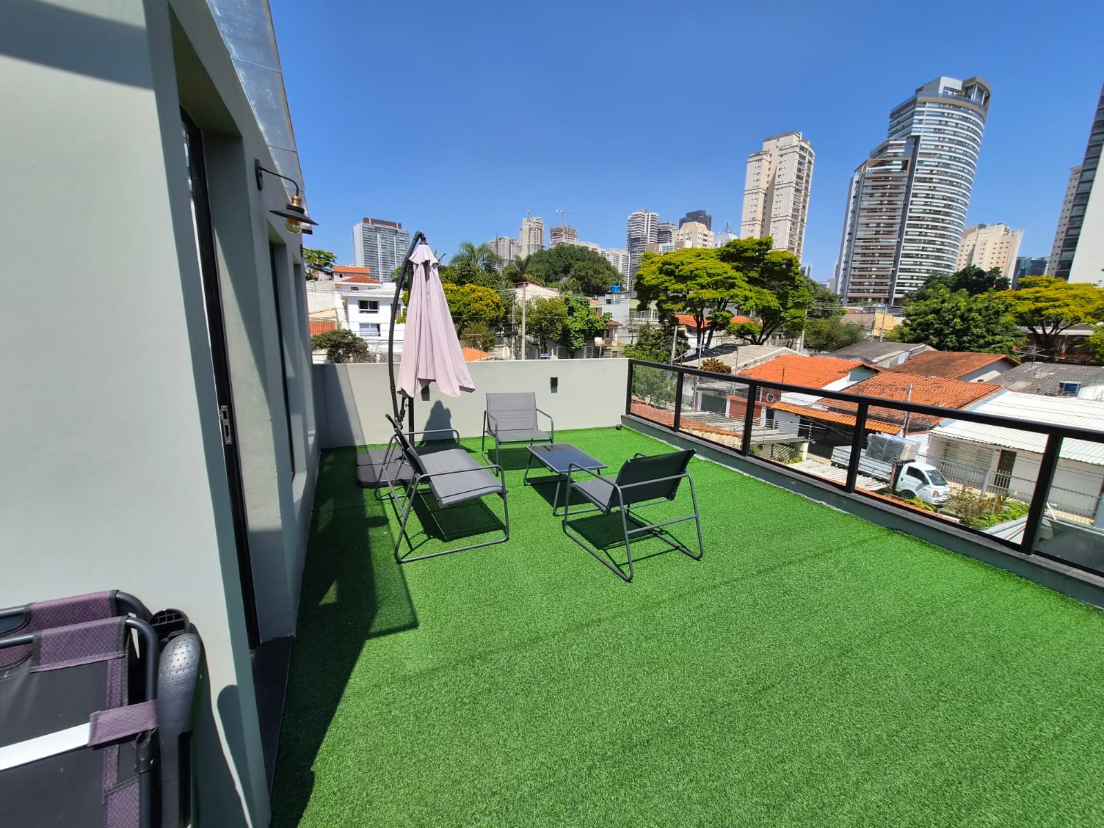 Terraço com grama sintética, guarda-sol e cadeiras, com prédios ao fundo